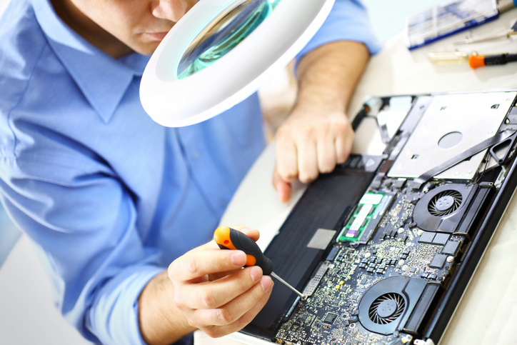 Unrecognizable adult caucasian man repairing a laptop. He's unscrewing tiny bolts on a motherboard. He's using magnifying glass with light. Visible cooling fans and RAM memory chips.